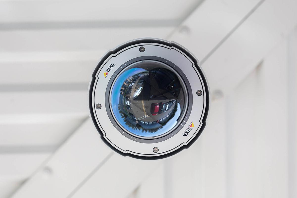 A close-up of a security camera against a white wall. 相机是安讯士品牌，在相机上可以看到停车场的倒影